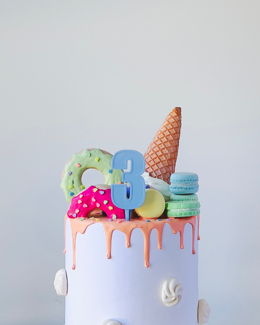 A colorful cake topped with a waffle cone, macarons, and sprinkles, featuring a large blue number three.