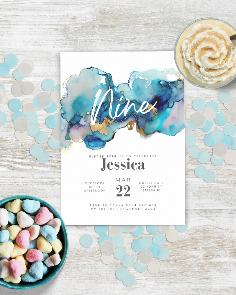 A colorful invitation featuring blue watercolor splashes, gold accents, and pastel heart-shaped candies on a rustic table.