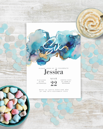 A colorful invitation featuring shades of blue and gold, surrounded by heart-shaped candies and confetti on a wooden table.