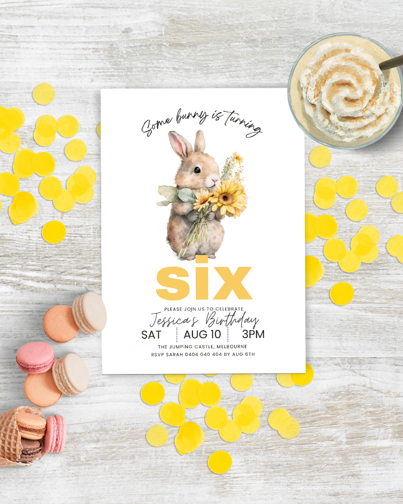 A cute bunny holds sunflowers on a birthday invitation with colorful confetti and treats on a wooden surface.