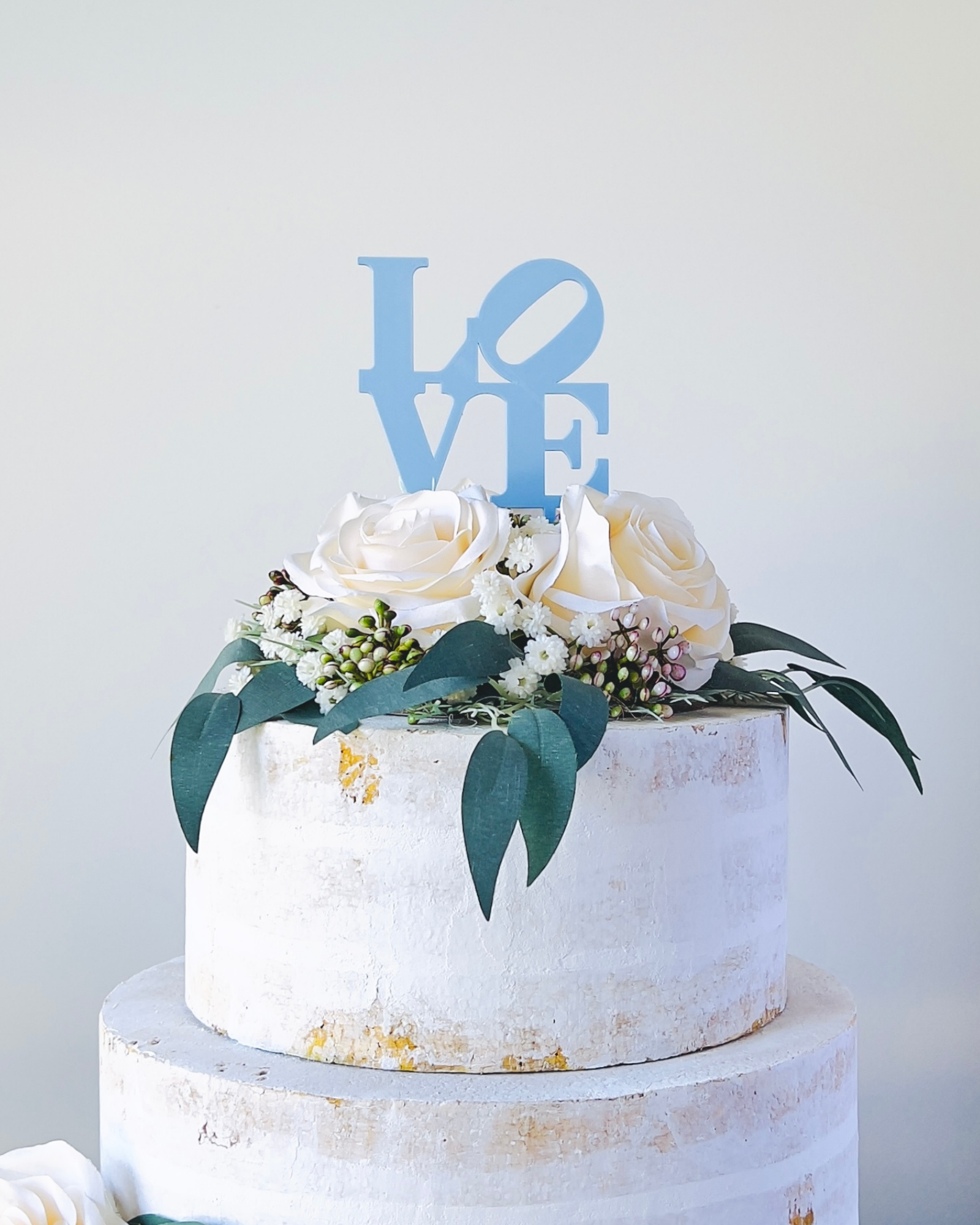 A tiered creation adorned with white roses, greenery, and a blue "LOVE" topper against a light backdrop.