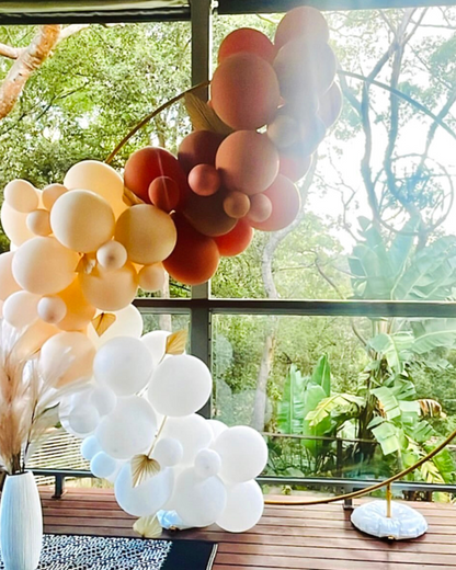 Colorful clusters of balloons in warm and cool tones, set against a lush green backdrop through large windows.