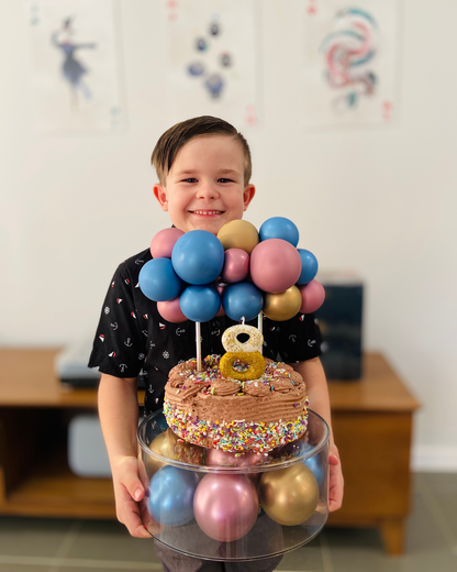 A young boy smiles while holding a colorful cake topped with balloons and a golden number 8, set against a bright background.