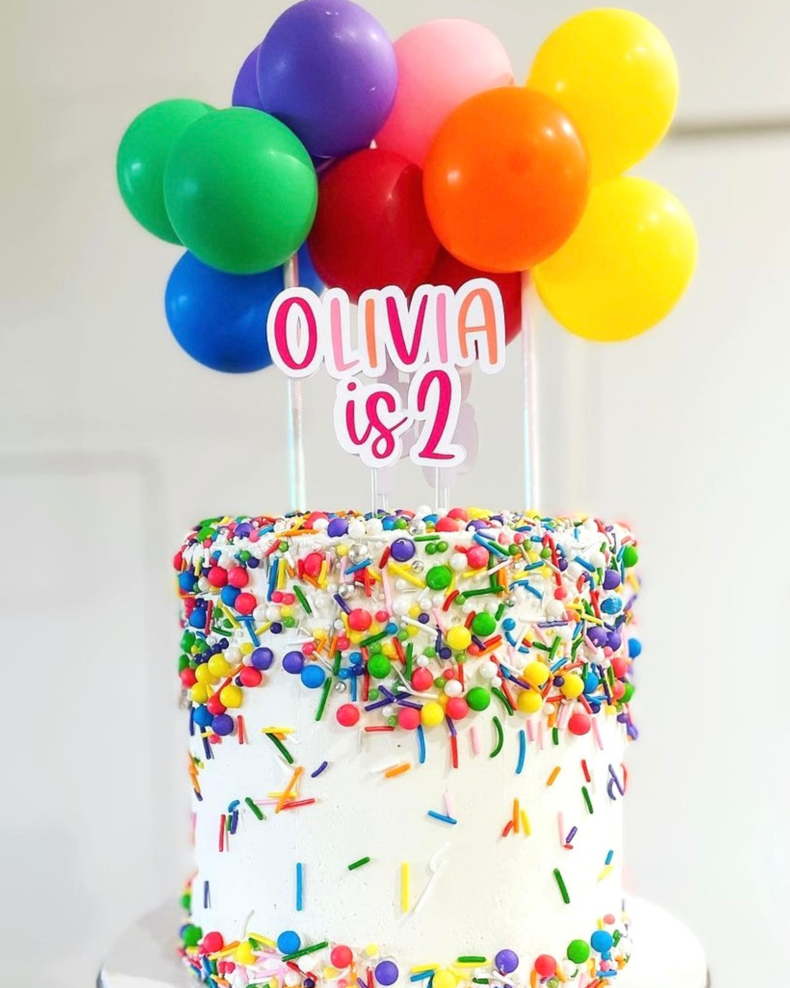 A festive cake topped with colorful balloons and a sign reading "OLIVIA is 2," adorned with vibrant sprinkles.