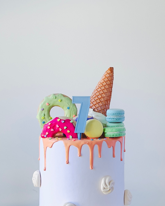 Colorful decorations atop a white cake, featuring playful elements like a cone, macarons, and a blue number seven.