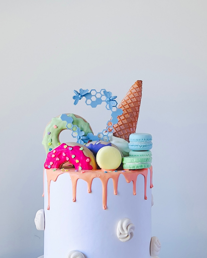 A colorful dessert topped with a cone, macarons, and donuts, featuring dripping icing and decorative bee elements.