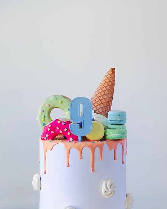 Colorful decorations atop a white cake, featuring a number 9, a cone, and vibrant sweets in playful arrangements.