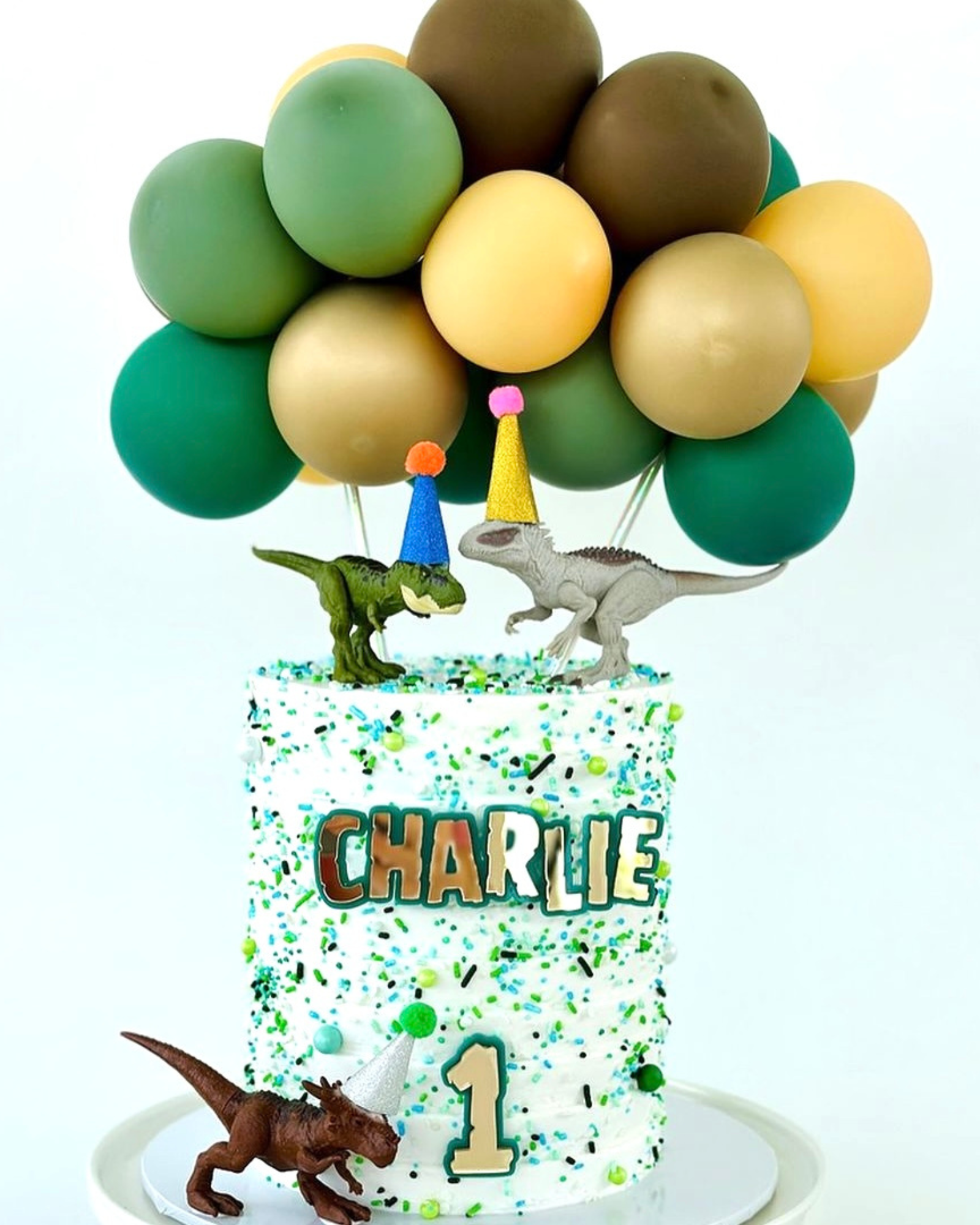 A colorful cake topped with dinosaur figures, festive balloons, and sprinkles, celebrating a first birthday.