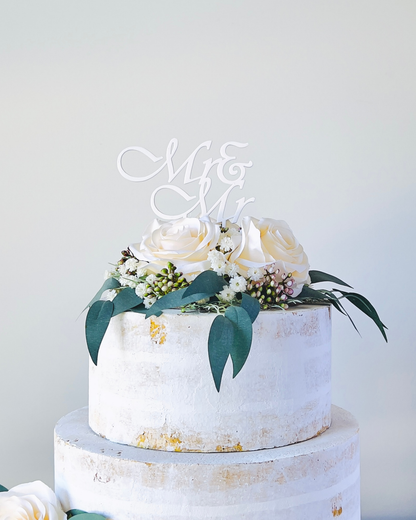 A two-tiered cake adorned with white roses, greenery, and a decorative "Mr & Mr" topper.