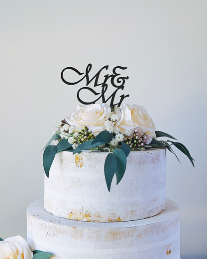 A three-tiered cake adorned with white roses and greenery, topped with a stylish "Mr & Mr" sign.