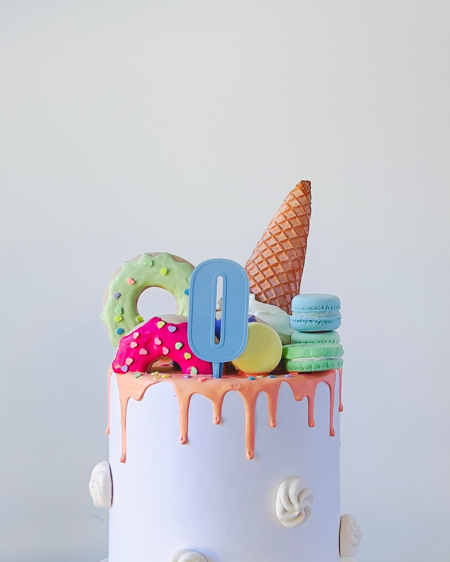 A colorful cake topped with playful treats, including a cone, macarons, and vibrant donuts, with a blue numeral zero.