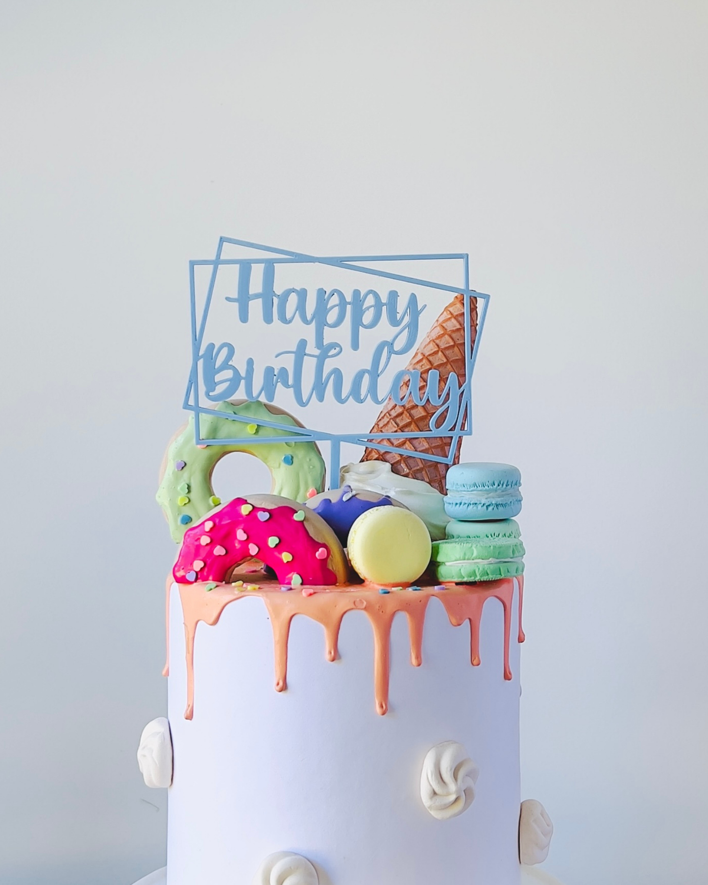 A colorful cake topped with donuts, macarons, and a cone, featuring a "Happy Birthday" sign in a light blue frame.