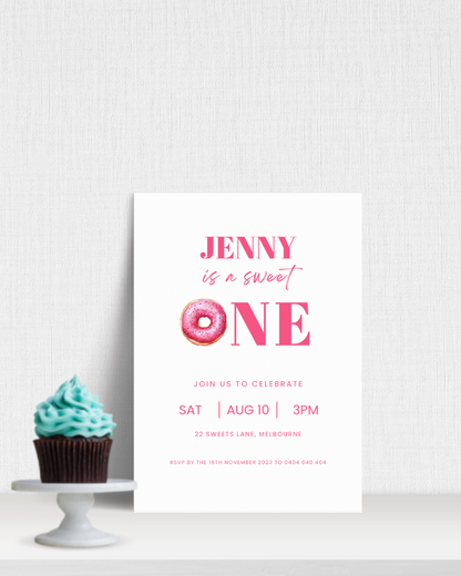 A cupcake with blue frosting beside a pink-themed invitation for a first birthday celebration.