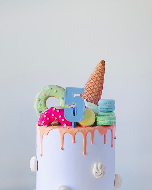 A colorful cake topped with a number 5, ice cream cone, macarons, and decorated sweets. Dripping frosting adds a fun touch.