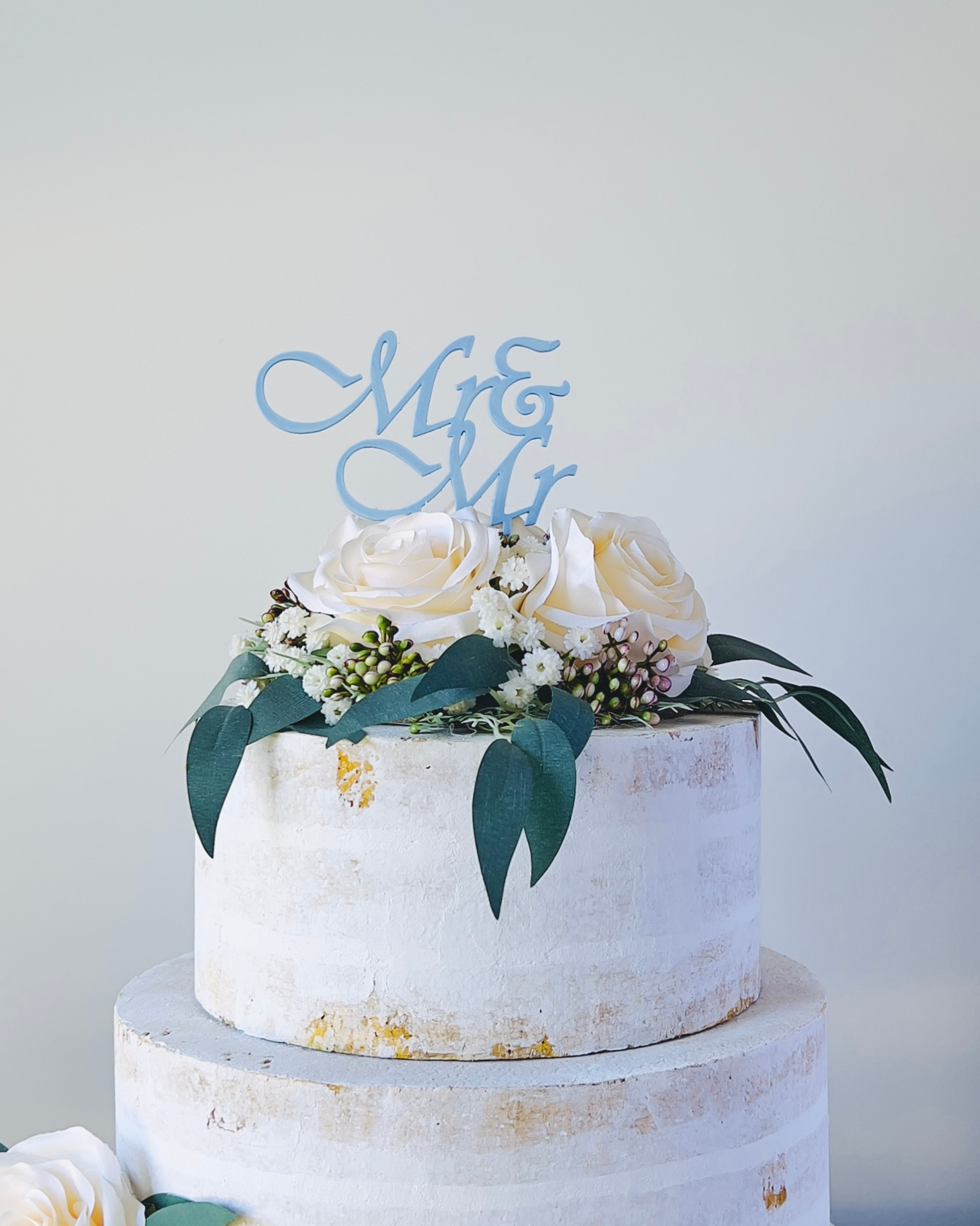 A three-tiered cake topped with white roses, greenery, and a blue "Mr & Mr" decoration.