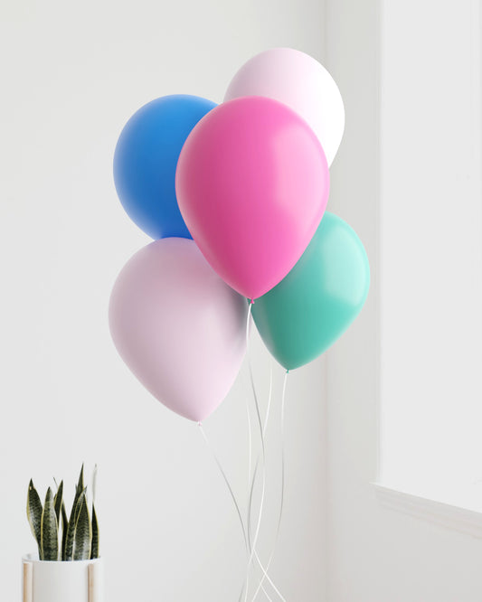 A cluster of colorful balloons in pink, blue, green, and white, floating above a potted plant against a bright wall.