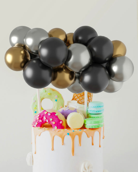 A festive arrangement of black, silver, and gold balloons atop a colorful cake decorated with sweets and drizzled frosting.