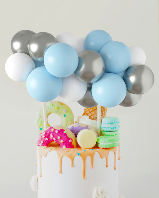 A colorful arrangement of blue, white, and silver balloons atop a cake adorned with vibrant sweets and dripping icing.