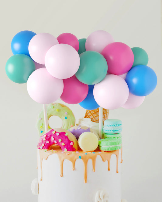 A colorful arrangement of balloons sits atop a cake decorated with vibrant sweets and dripping icing.