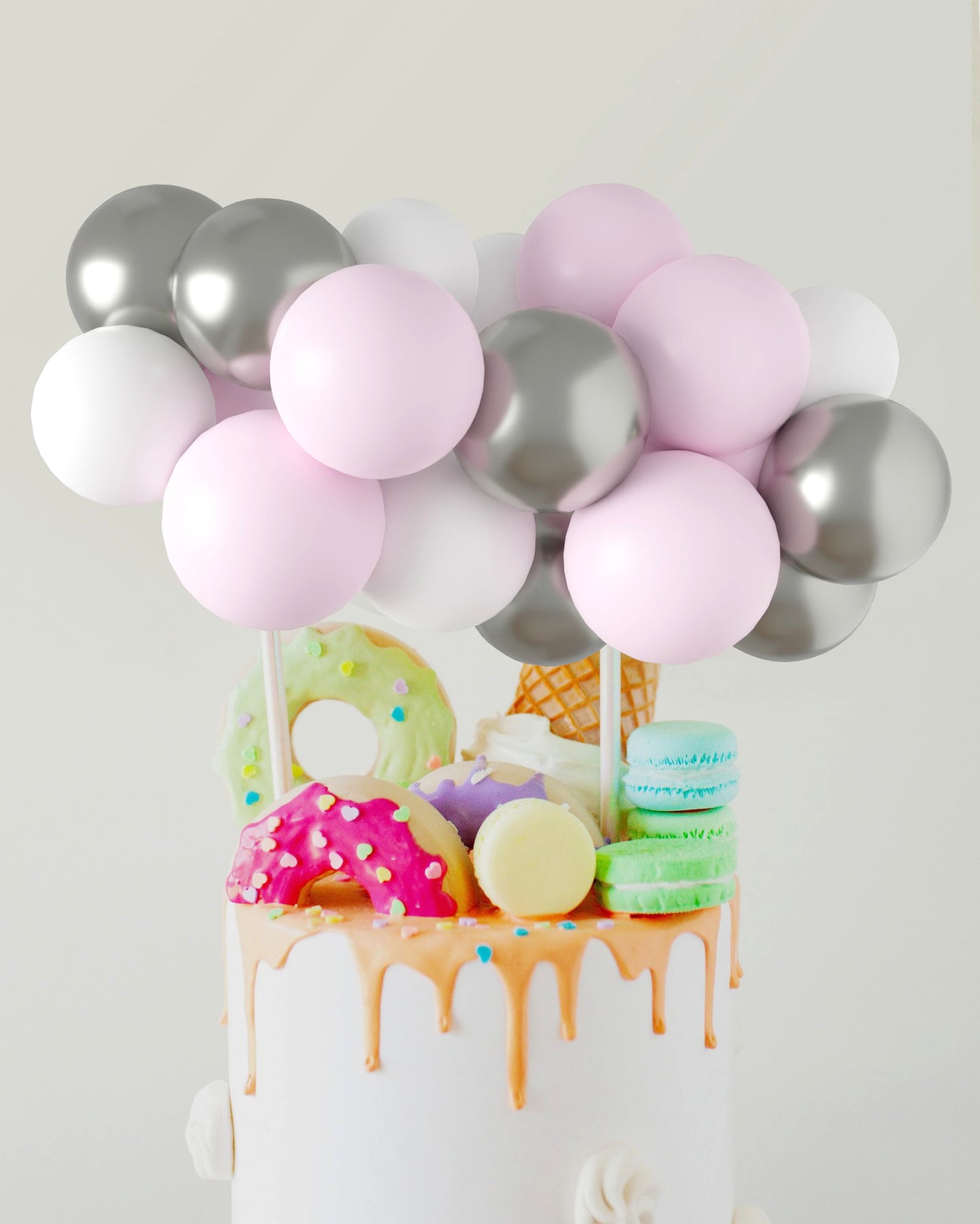 A colorful arrangement of pink, white, and silver balloons atop a decorated cake featuring donuts and macarons.