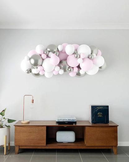 A colorful balloon garland in pink, white, and silver decorates a wall above a modern wooden table and lamp.