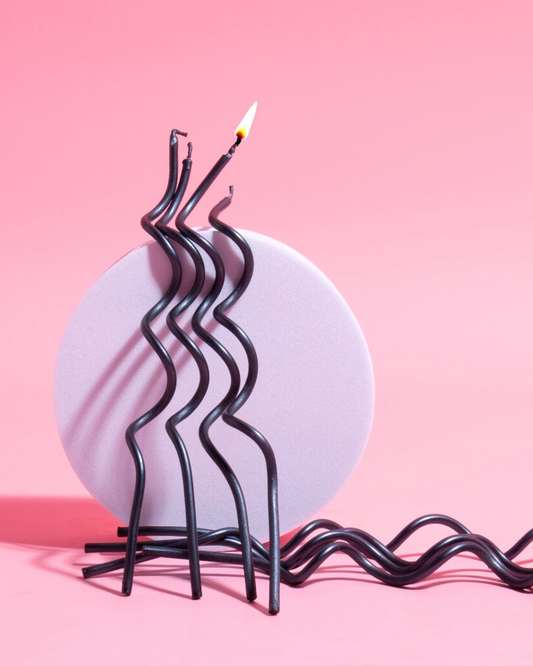 Curvy black candles with a flame stand on a round lavender base against a pink backdrop, creating a playful composition.