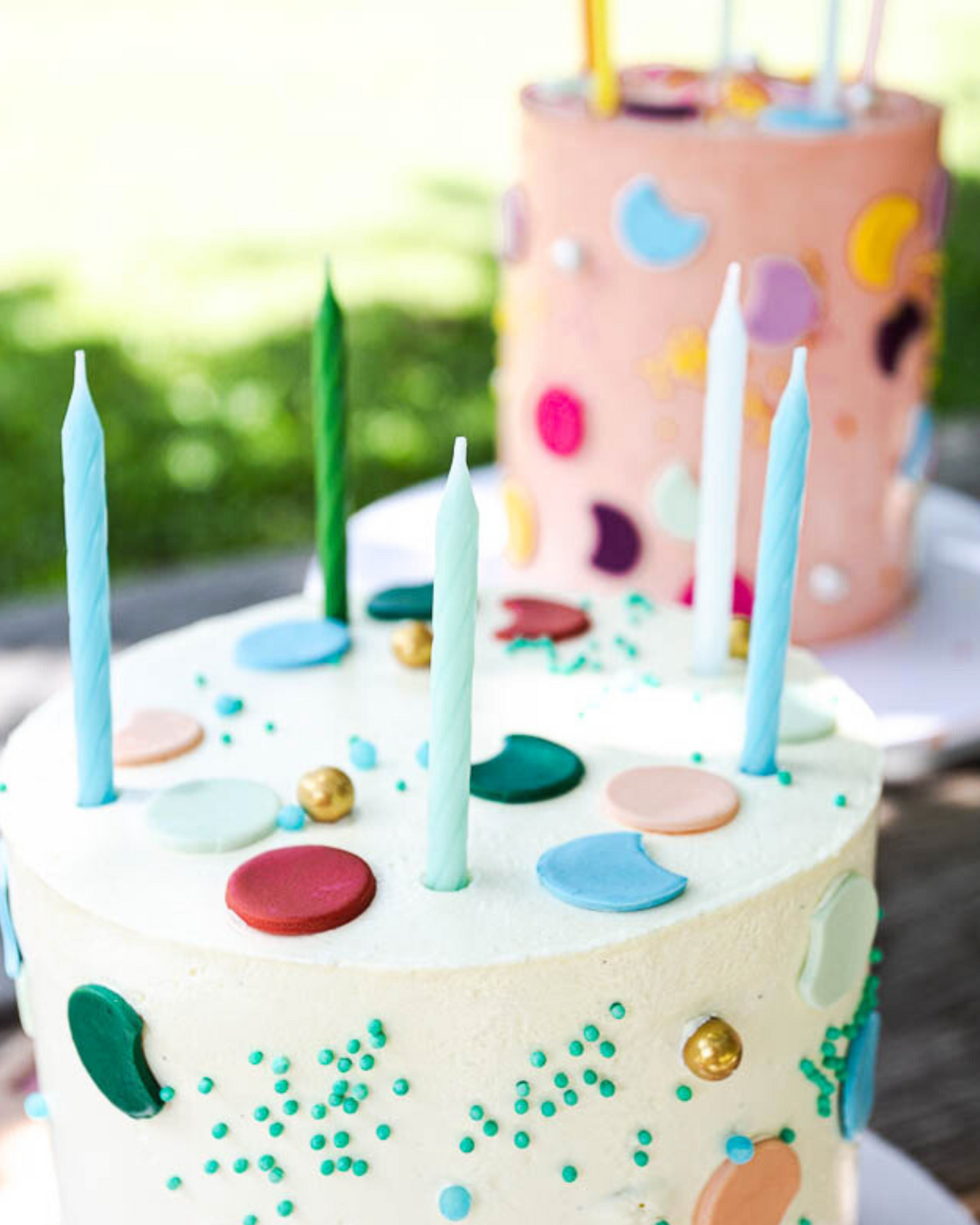 Two colorful cakes with candles, adorned with vibrant dots and sprinkles, set against a blurred green backdrop.