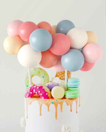 A whimsical cake topped with pastel balloons and colorful treats like donuts, ice cream, and macarons.