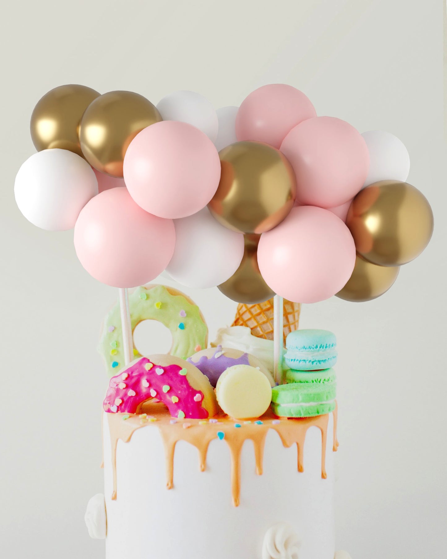 A festive arrangement of pink, white, and gold balloons atop a whimsical cake adorned with colorful treats.