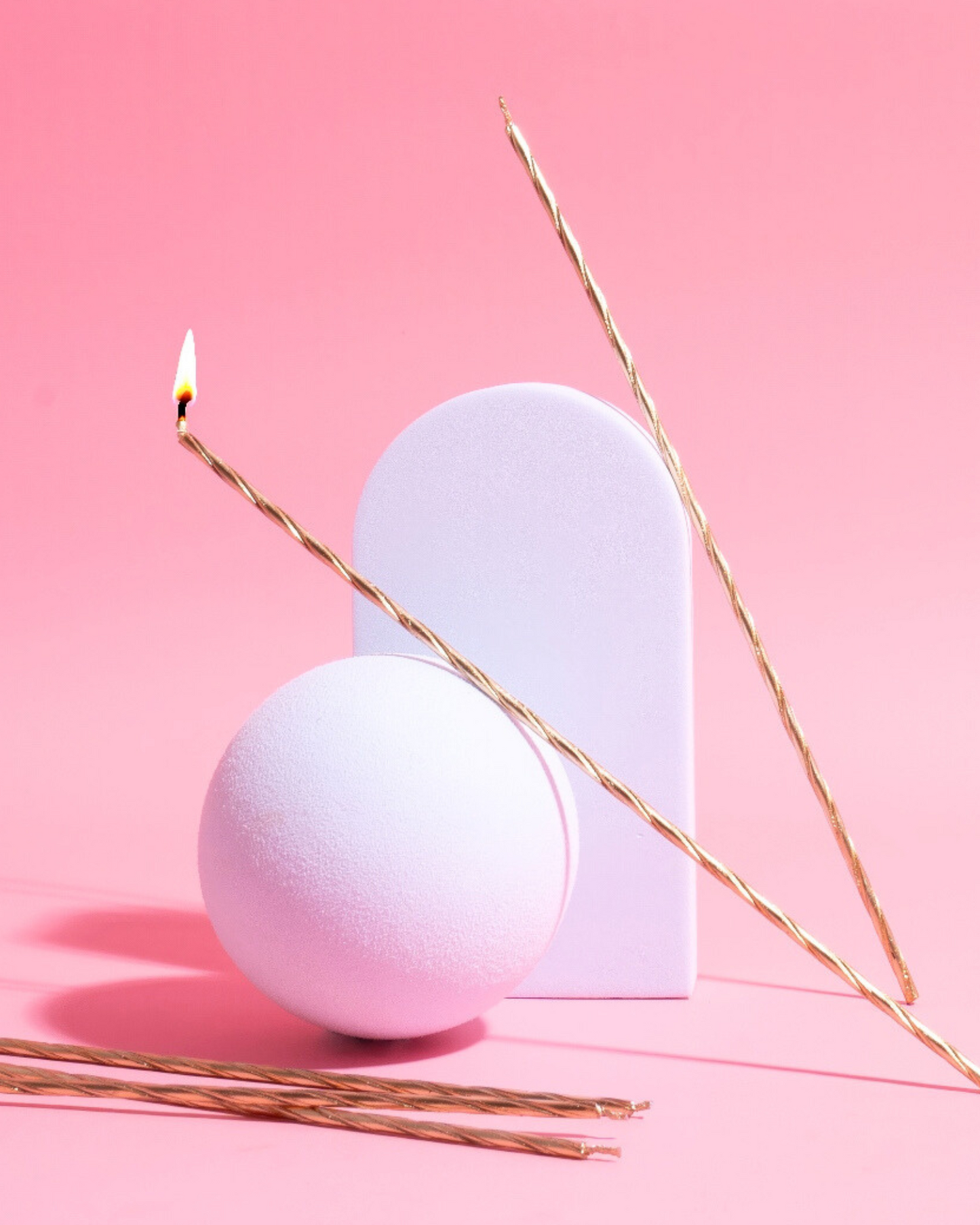 Gold spiral candles and a round object are arranged against a soft pink background, creating a modern aesthetic.