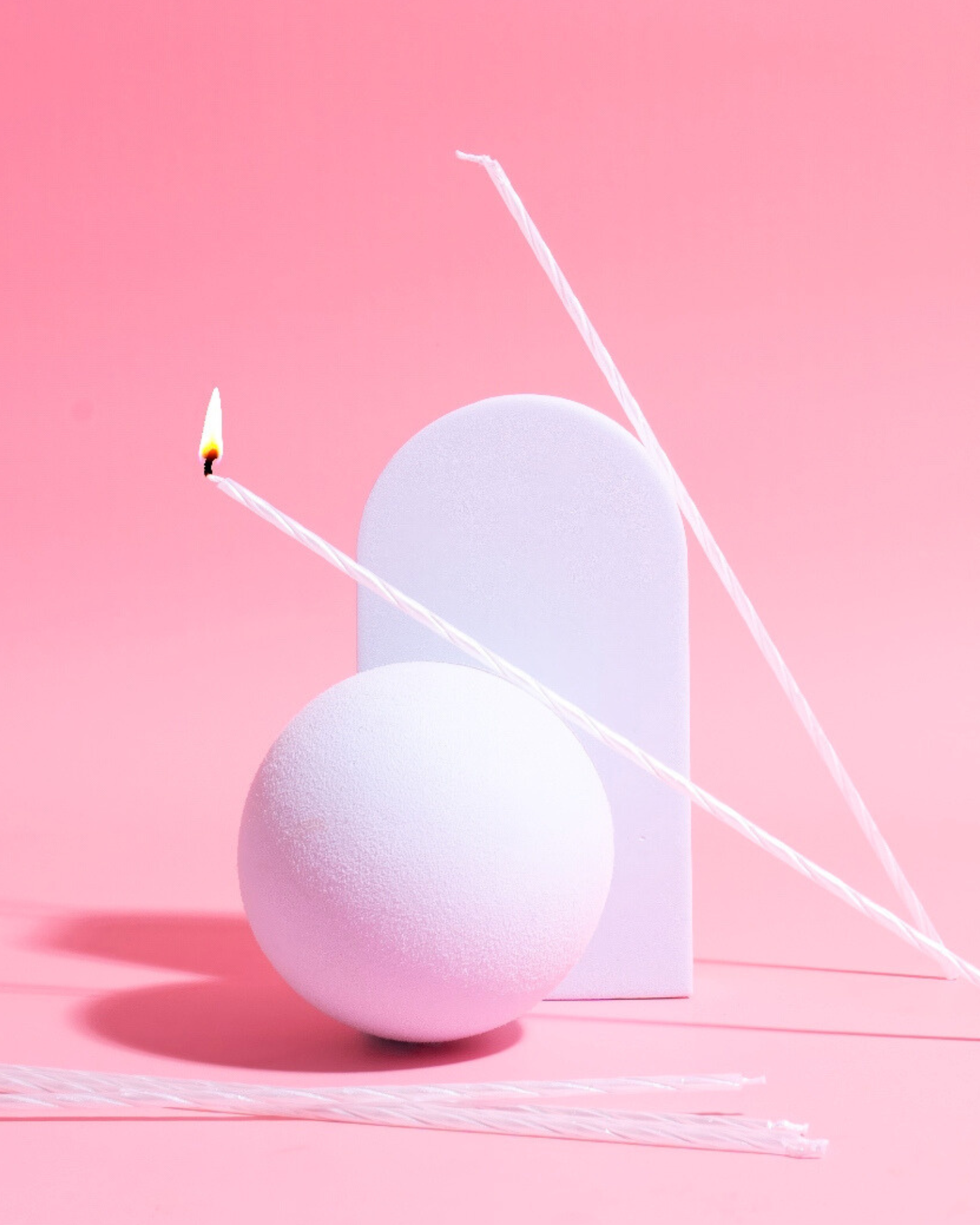 A minimalist arrangement featuring white spiral candles, a round object, and a rectangular form against a soft pink backdrop.