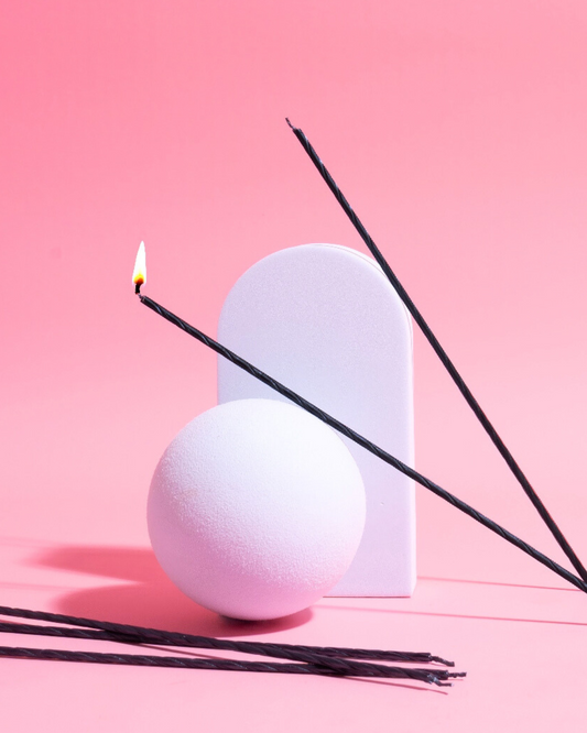 Black spiral candles with flames positioned beside a textured white sphere and an arch against a soft pink background.