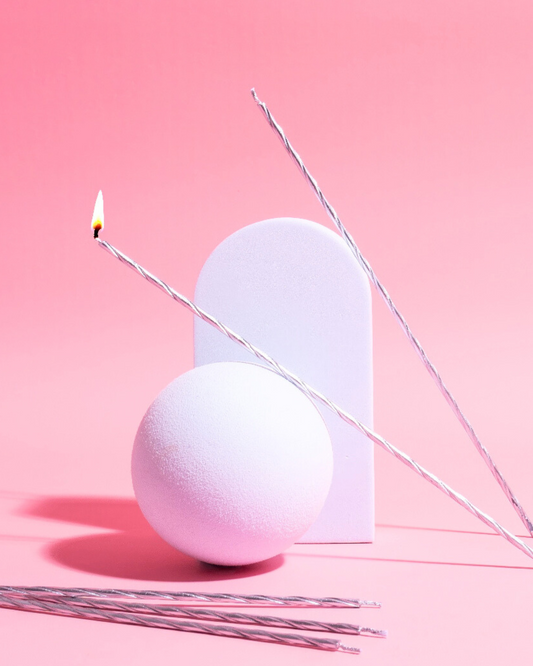 A soft pink background features a round white sphere, a rectangular shape, and twisting silver candles, one lit.