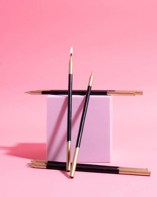 Black cake candles with gold tips arranged around a pastel pink cube against a bright pink background.