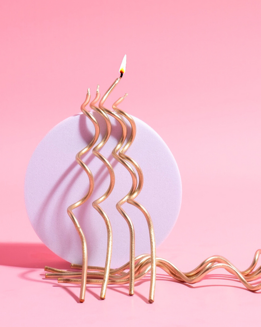 Three swirling gold candles burning atop a soft purple round base against a vibrant pink background.