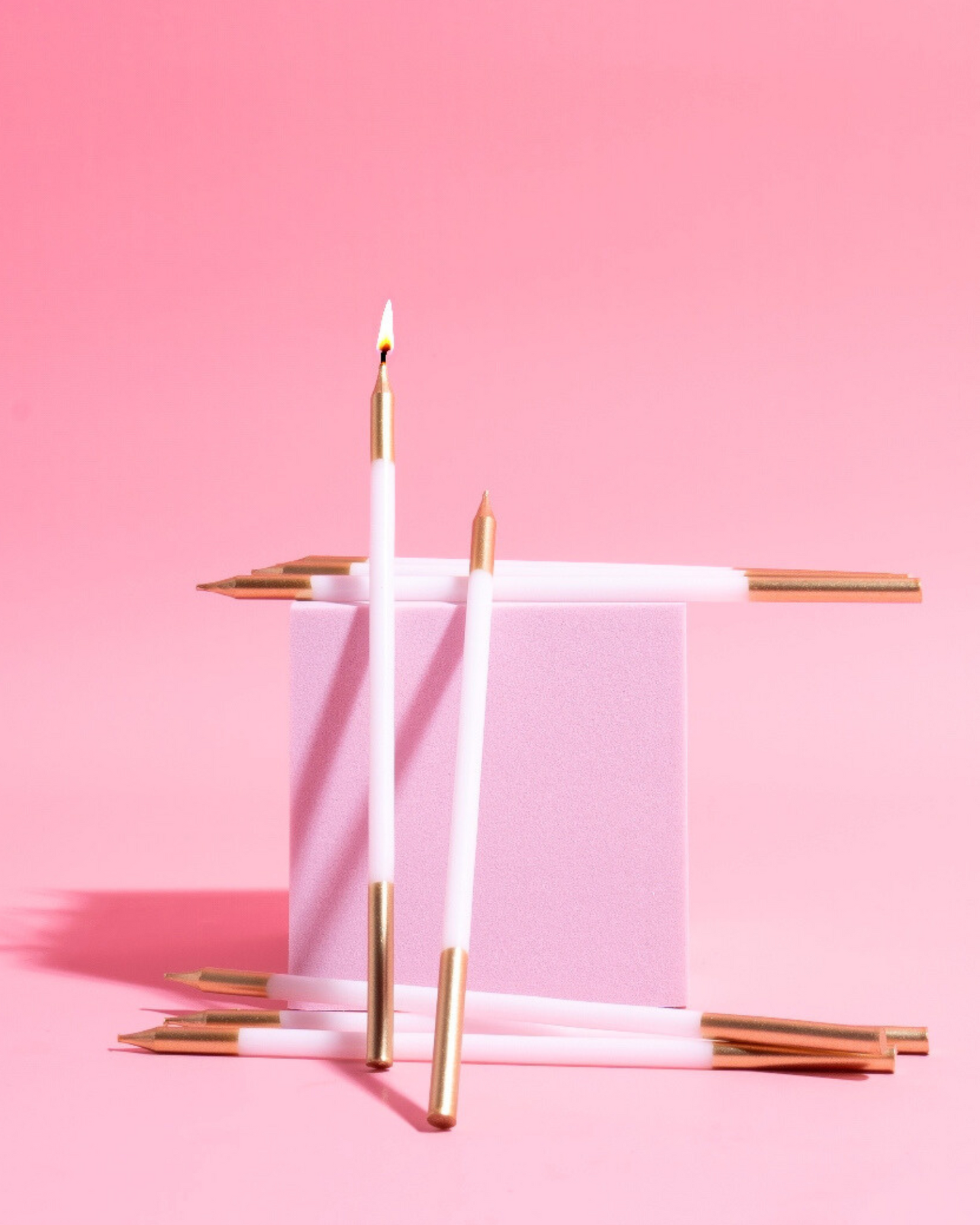 White candles with gold tips arranged around a soft pink block on a pastel pink background.