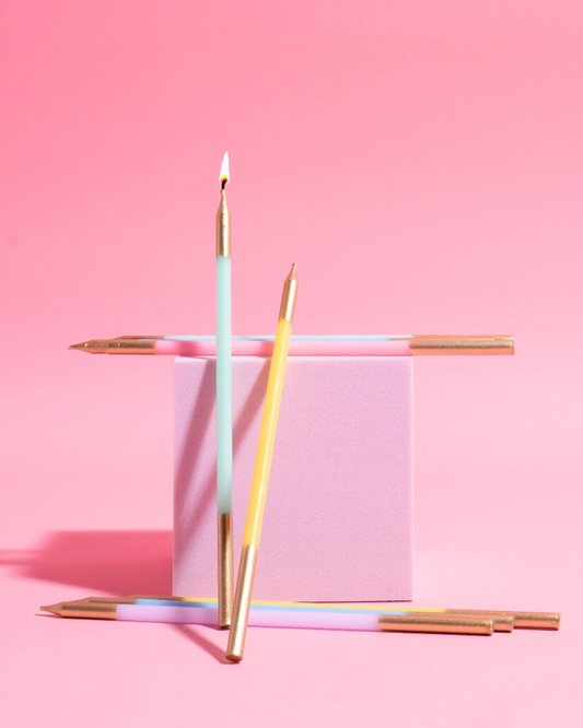 Colorful pastel candles with gold tops arranged around a soft pink block against a vibrant pink background.