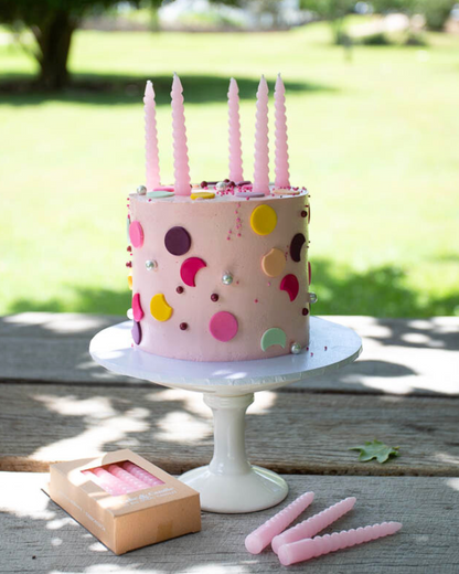 A pink frosted cake with colorful sprinkles and five pink candles, set on a white pedestal outdoors.