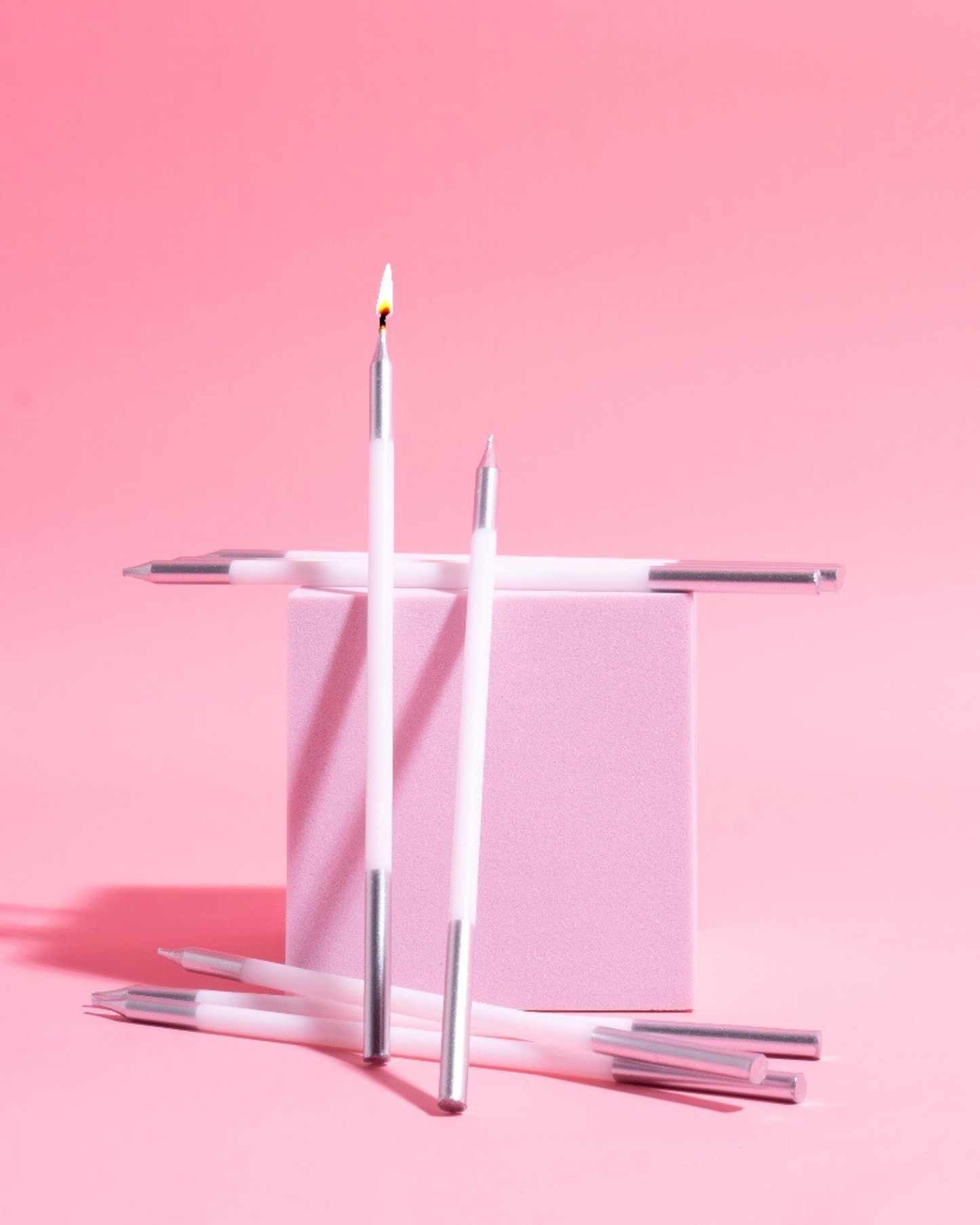 White candles with silver tips arranged on a soft pink background, one candle lit, creating a festive atmosphere.