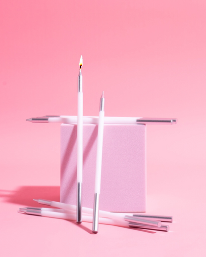 White candles with silver tips arranged on a soft pink background, one candle lit, creating a festive atmosphere.