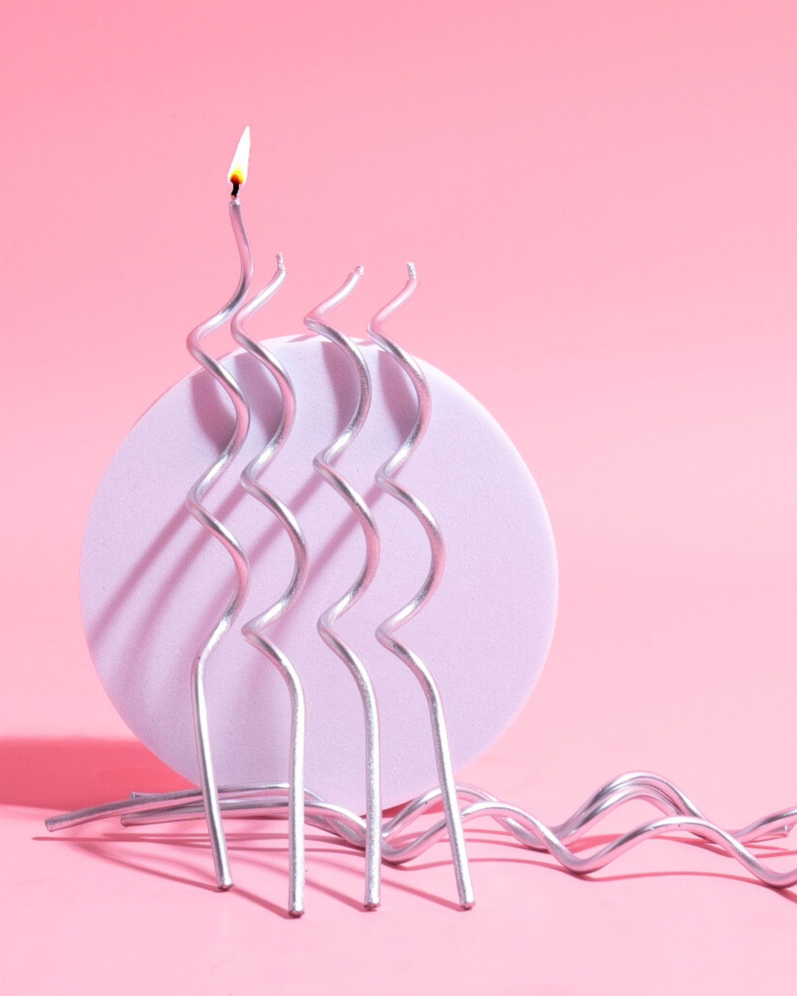 Silver, wavy candles stand on a round pastel base, against a soft pink background, with one candle lit.