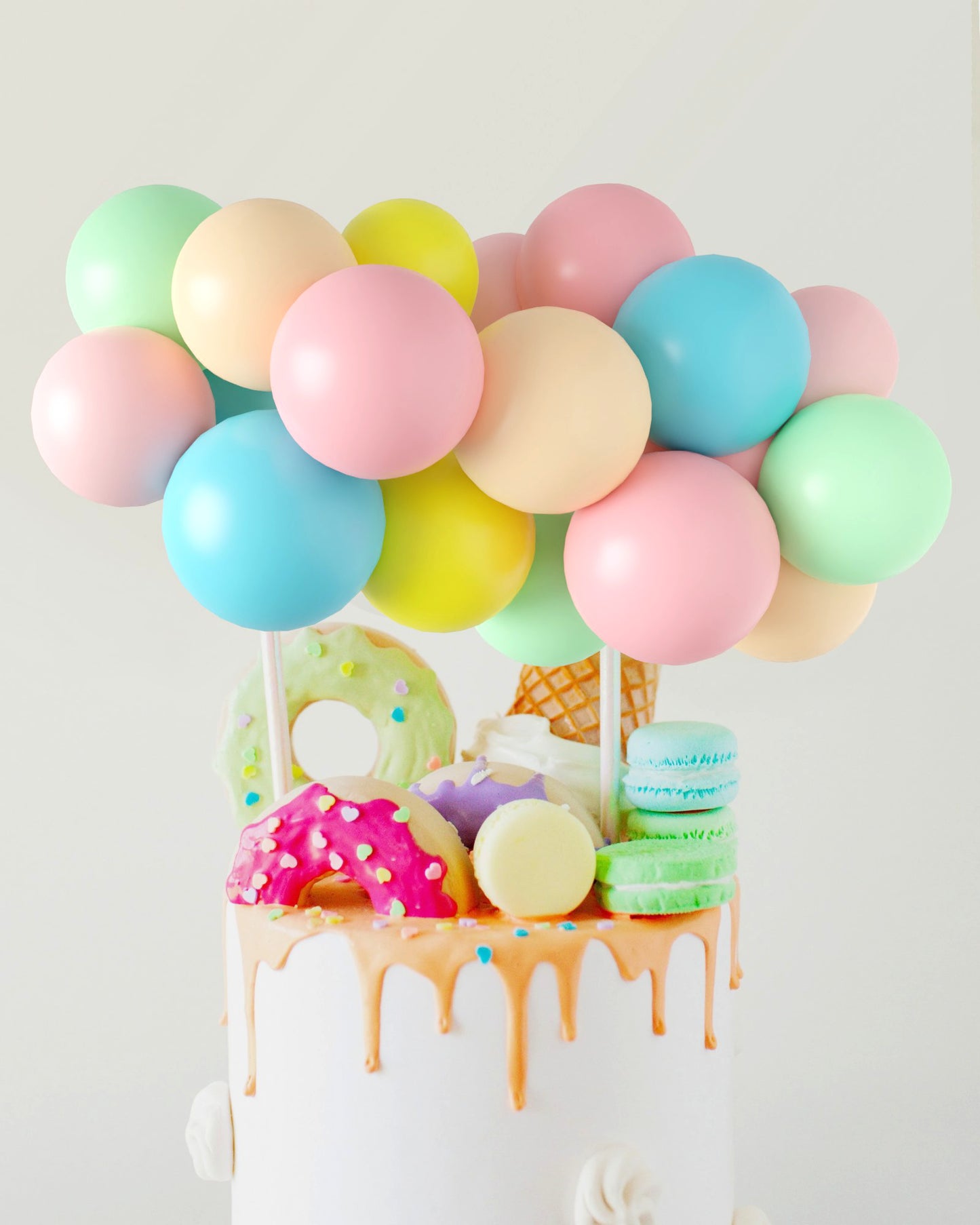 Colorful balloons hover above a cake adorned with pastel treats, including donuts, macarons, and an ice cream cone.