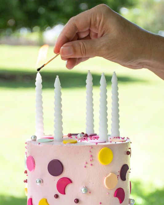 A hand lights a white spiral candle atop a pink cake adorned with colorful sprinkles and decorations in a sunny outdoor setti