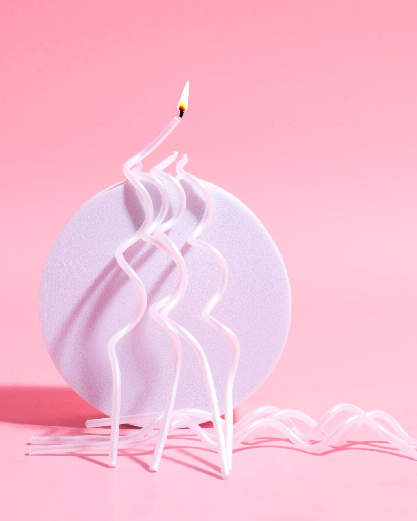 Curvy white candles with a flame stand on a round lavender base against a soft pink background.