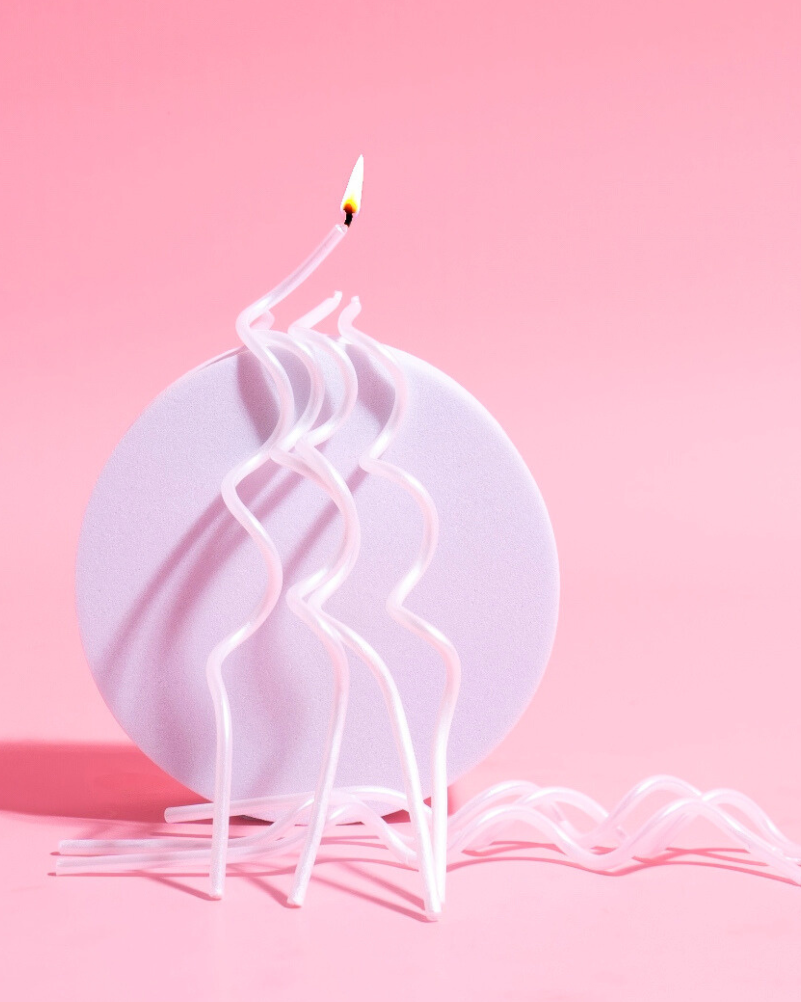 Curvy white candles with a flame stand on a round lavender base against a soft pink background.