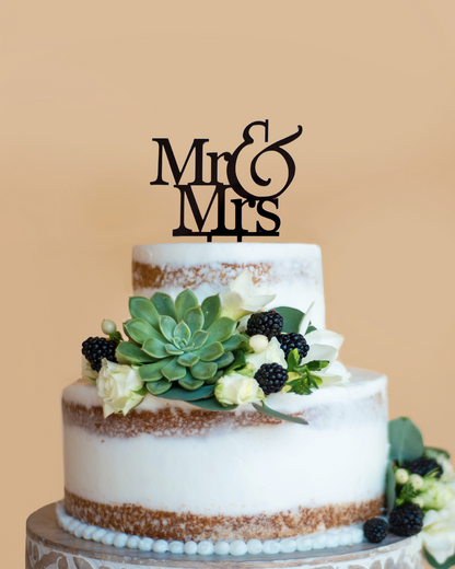 Three-tiered cake adorned with greenery, blackberries, and a stylish "Mr & Mrs" topper against a soft background.