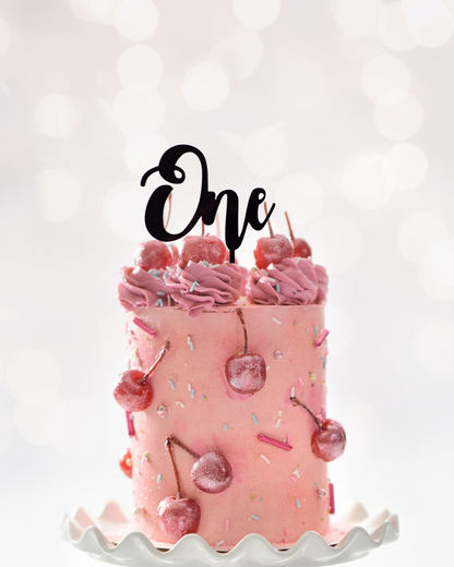 A pink cake topped with swirls and cherries, featuring a stylish black "One" topper against a soft, blurred background.