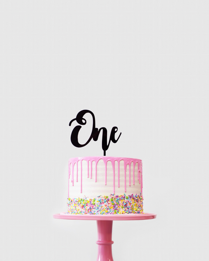 A pink-frosted cake with colorful sprinkles topped with a black "One" topper on a pink pedestal.