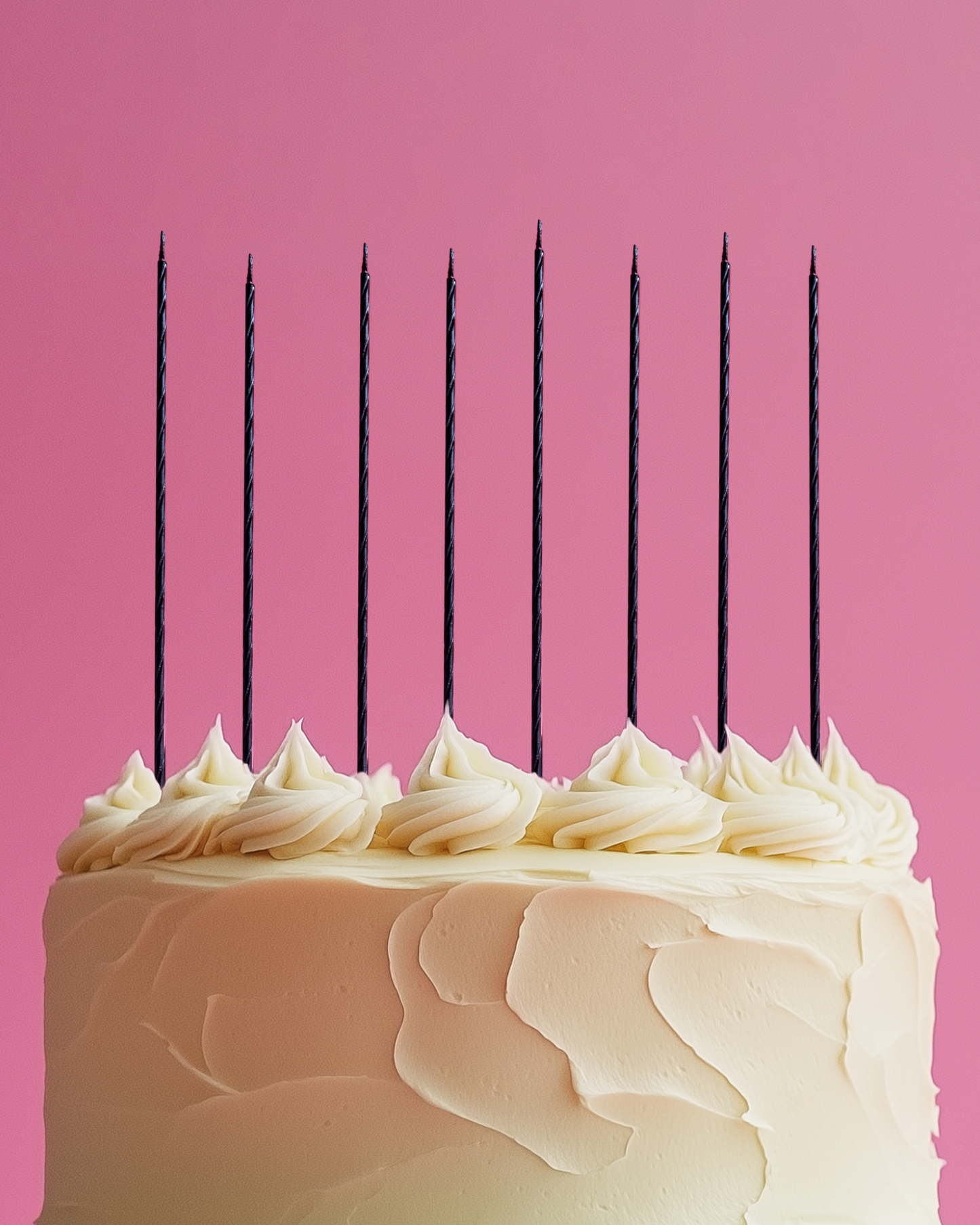 A tall cake with smooth frosting and dark candles stands against a pink background, creating a festive atmosphere.