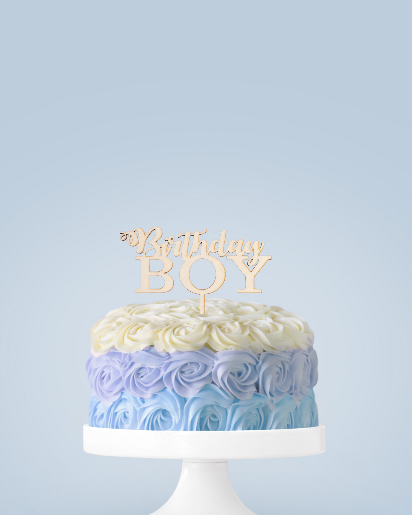 A beautifully piped cake with blue ombre rosettes and a wooden "Birthday Boy" topper, set against a light background.